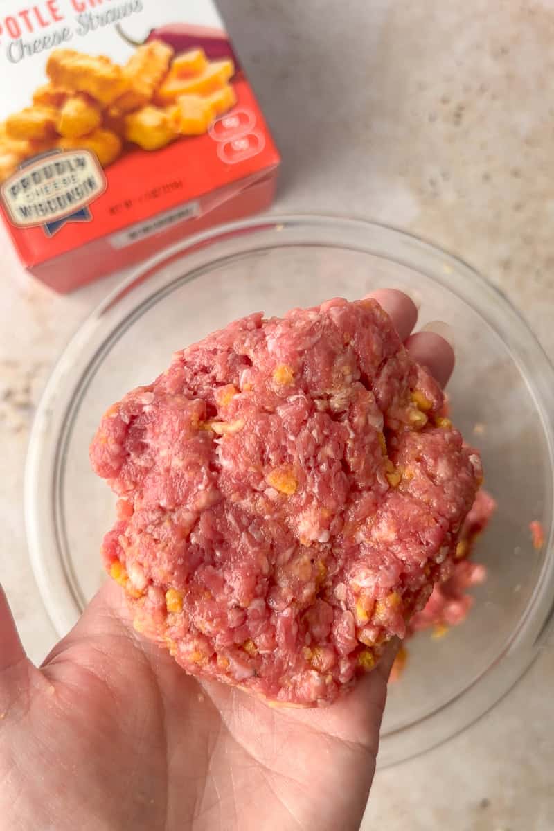 Formed raw burger patty held in a hand, showing bits of crushed cheese straws mixed throughout.