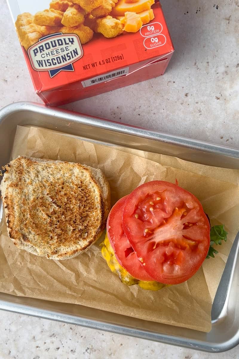 Assembled burger with cheddar, lettuce, and tomato on a toasted bun, sitting open-faced on a tray next to Mama Geraldine's box.