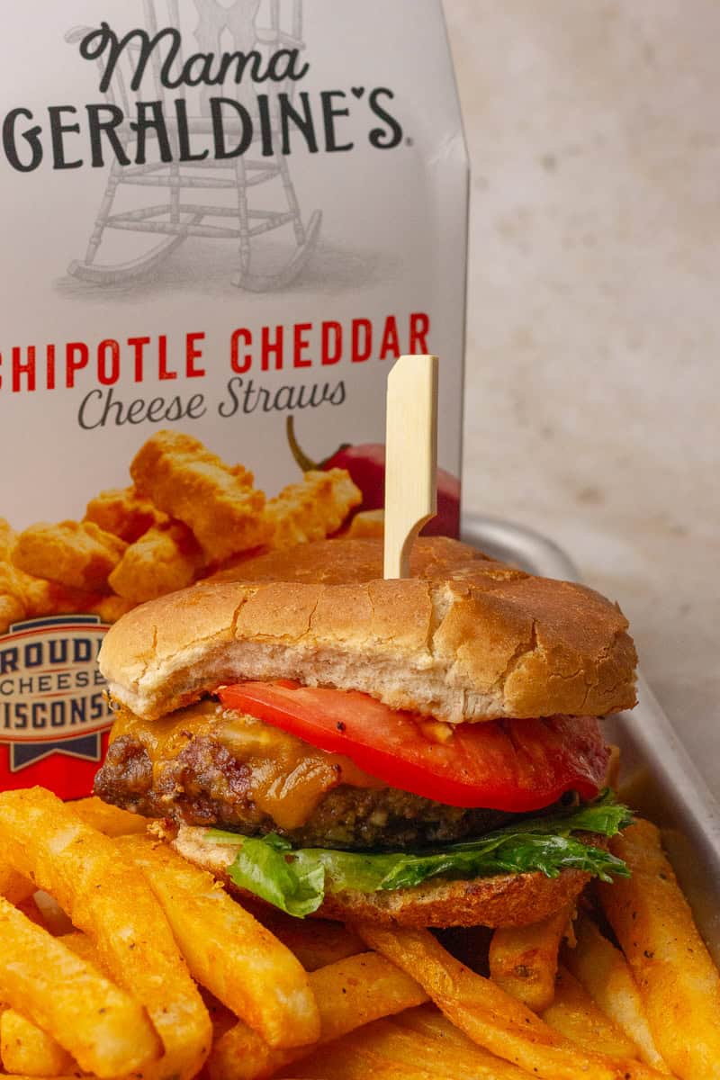 Close-up of a grilled chipotle cheese burger served with fries and Mama Geraldine’s cheese straws in the background.