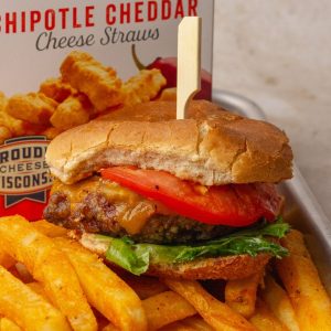 Grilled burger with cheddar, lettuce, and tomato on a toasted bun, placed in front of a Mama Geraldine's cheese straws bag.