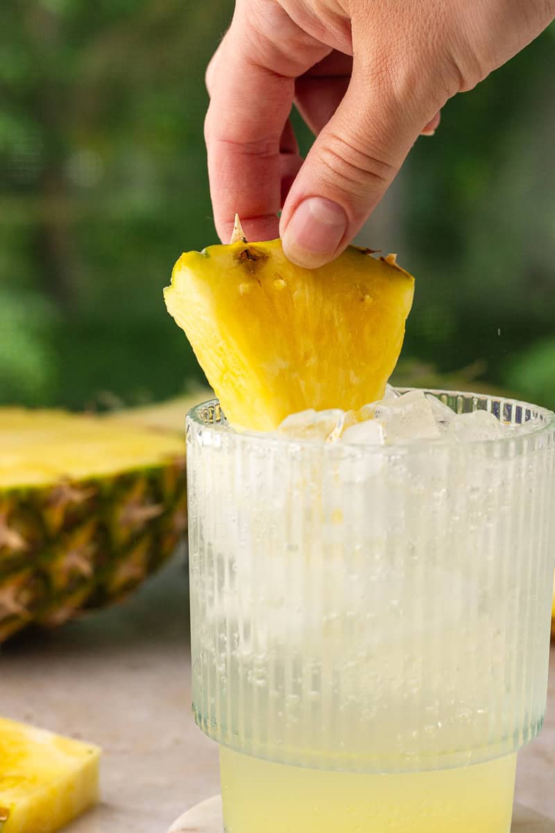 A hand placing a pineapple wedge garnish into a finished glass of pineapple ranch water.