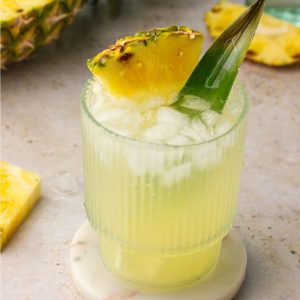 A styled shot of pineapple ranch water on a light surface, garnished with pineapple and surrounded by fresh fruit.