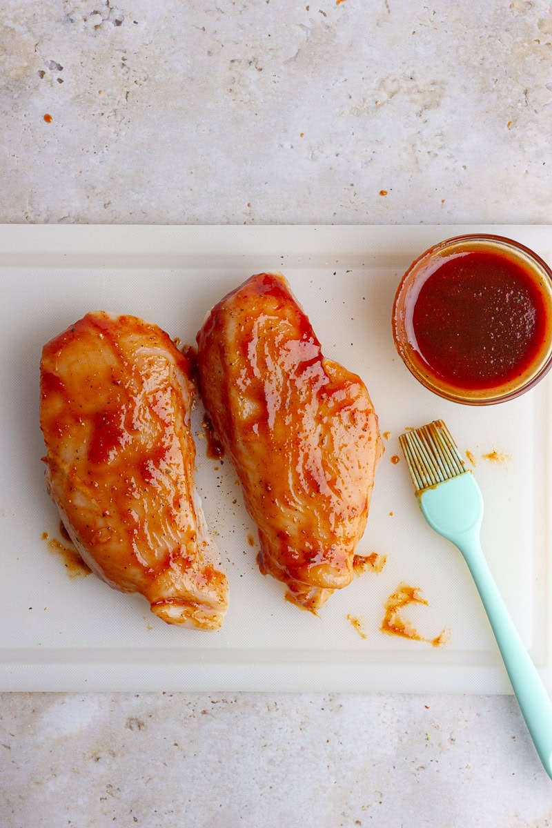 Chicken breasts brushed with BBQ sauce, prepped on a cutting board next to a small bowl of sauce and basting brush.