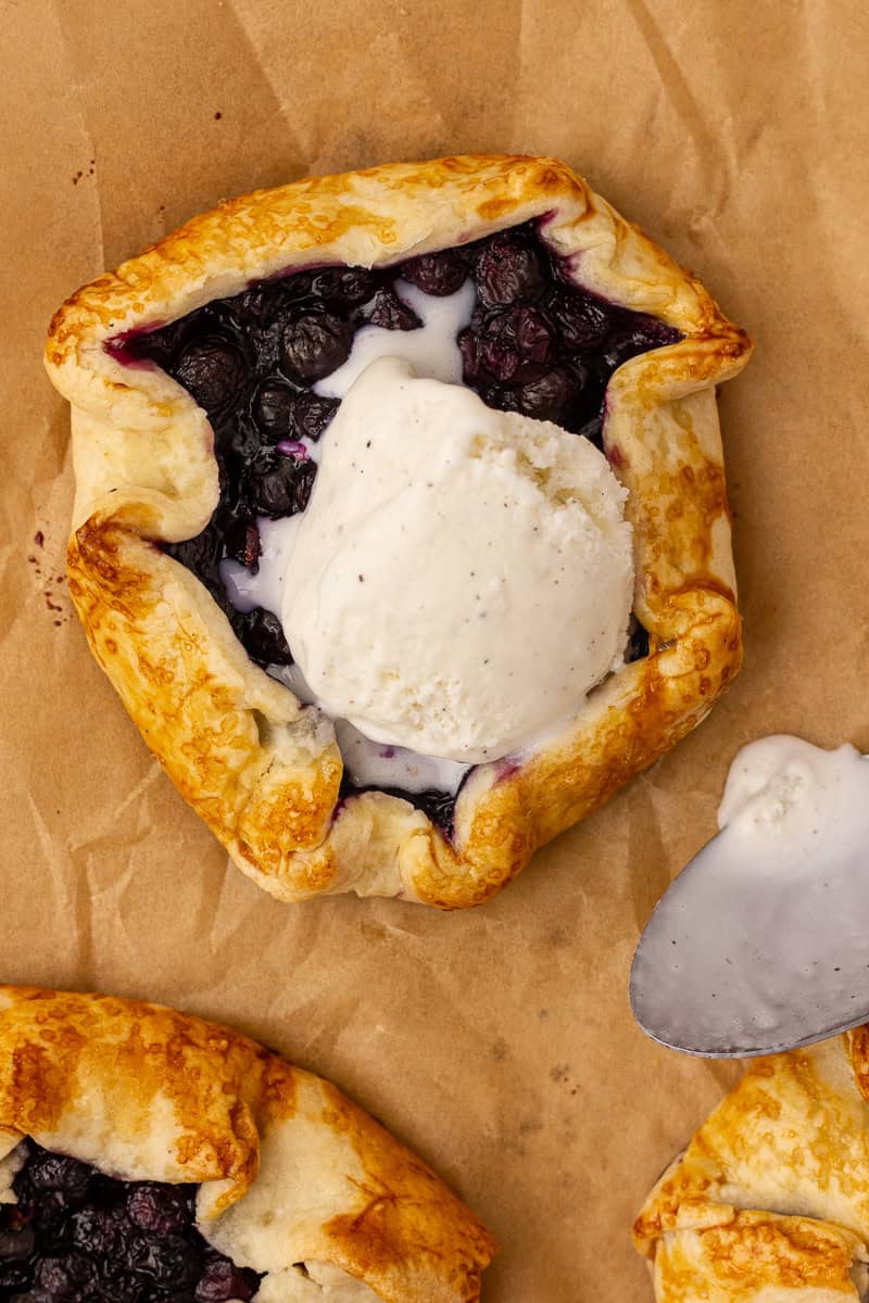 A mini blueberry lemon galette topped with melting ice cream sits beside a spoon and a drip of melted cream.