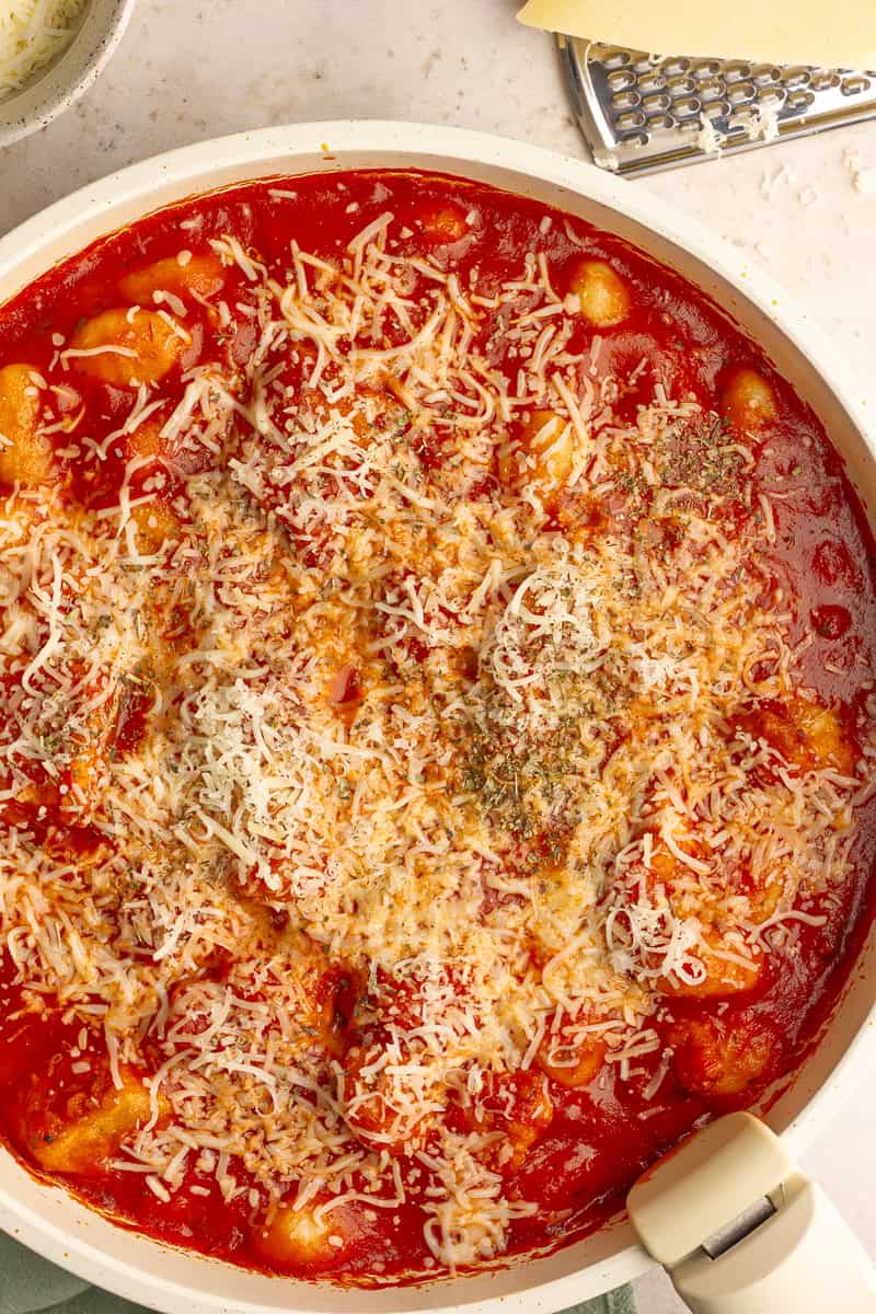 Gnocchi simmering in marinara sauce topped with shredded cheese in a skillet.