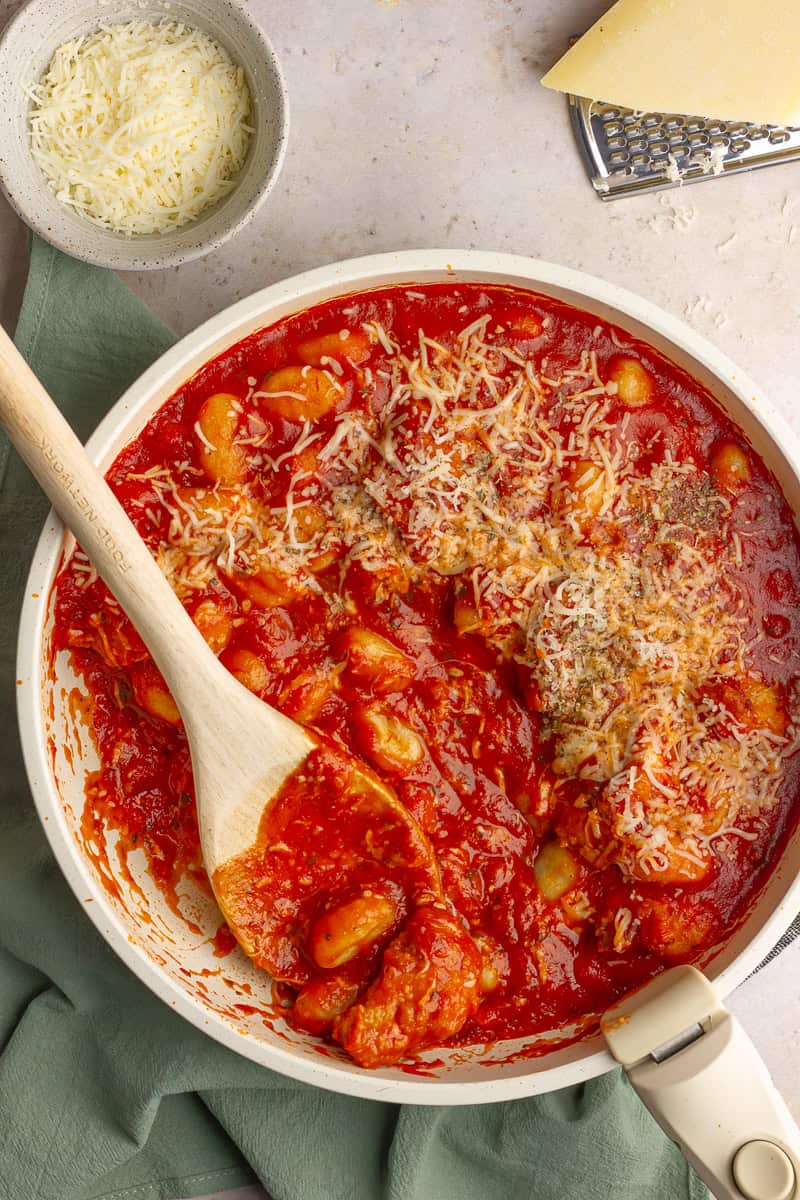 A wooden scooping stirring cheesy gnocchi and marinara in a white skillet.
