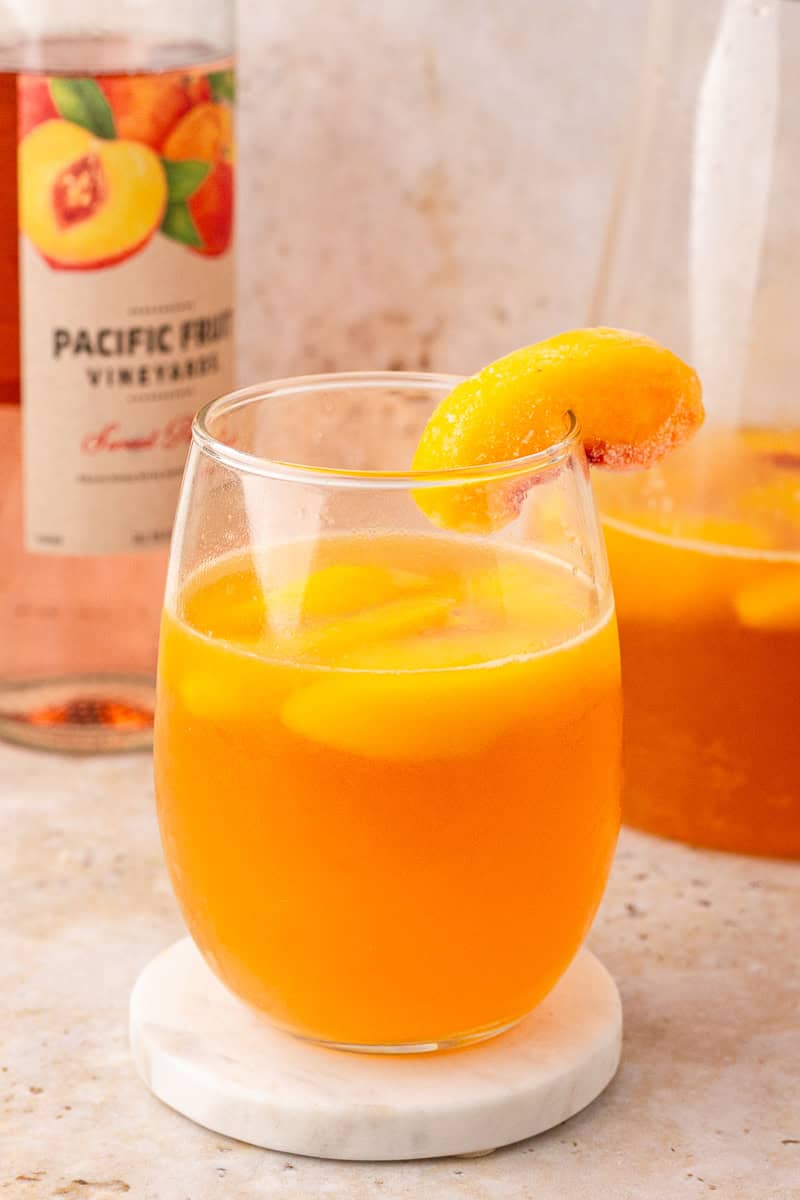 A bright and refreshing glass of peach sangria with a frozen peach slice on the rim, pitcher in the background.