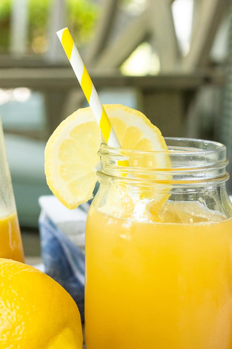 A close-up of a mason jar filled with peach lemonade, garnished with a lemon slice and yellow-striped straw.