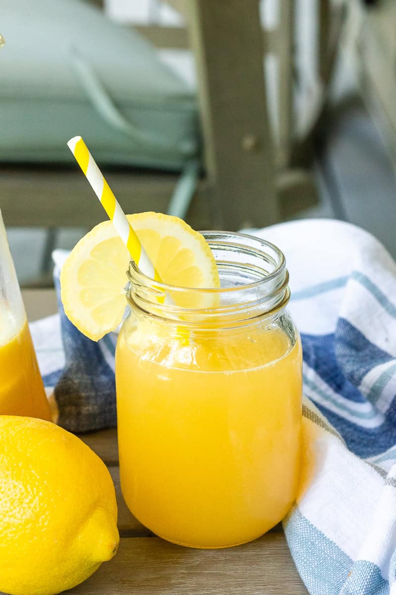 A full view of a glass of southern peach lemonade on a porch table next to fresh lemons and a tea towel.