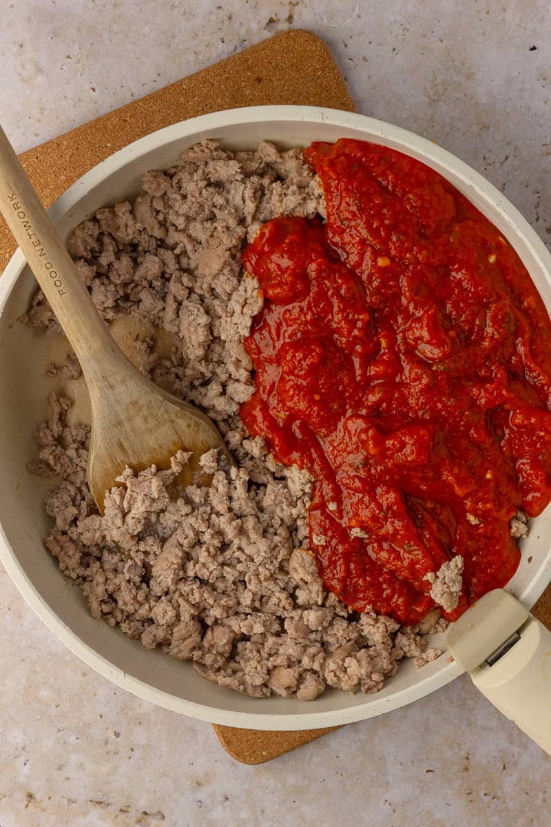 A skillet with browned ground turkey and marinara sauce being stirred together with a wooden spoon.