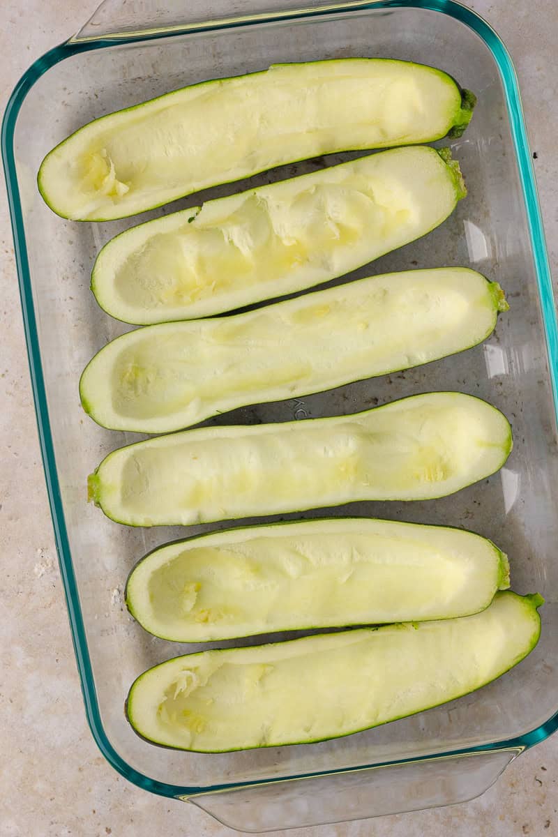 Six halved zucchinis with the centers scooped out, arranged in a glass baking dish.