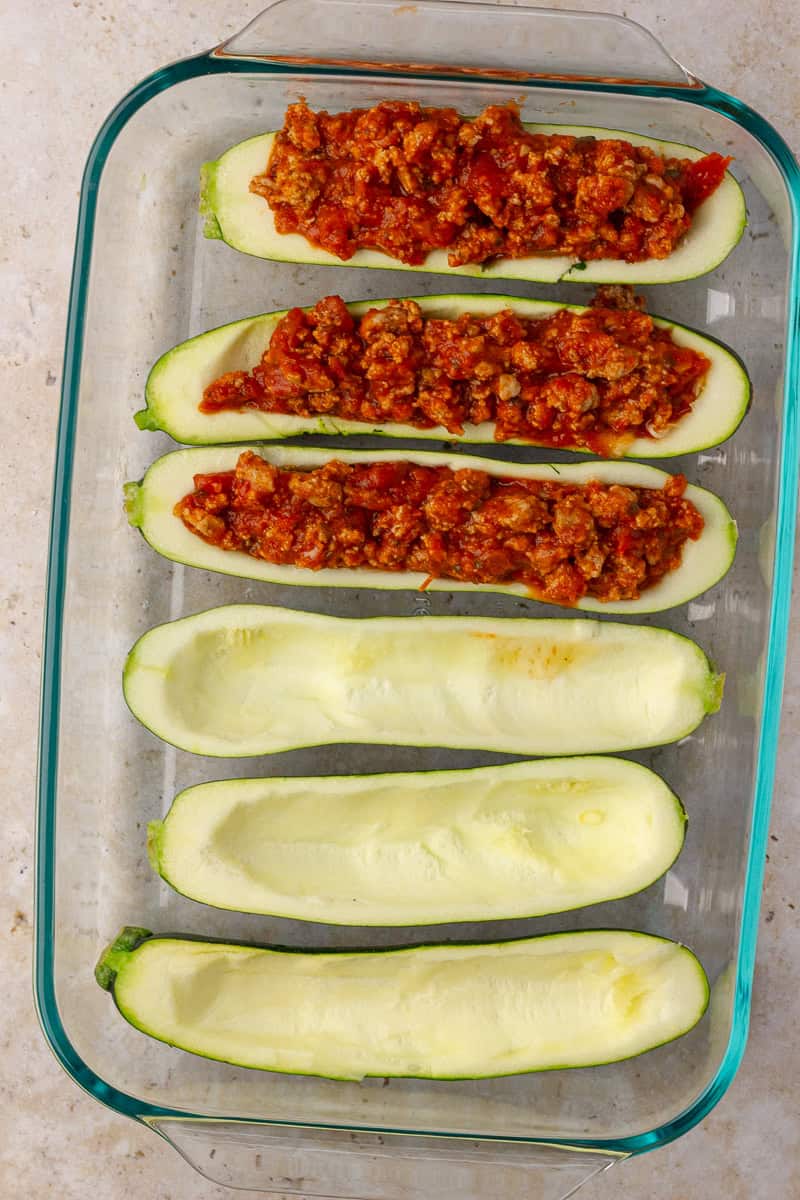 Four zucchini boats filled with bolognese mixture in the top row of a baking dish, with two empty boats on the bottom row.