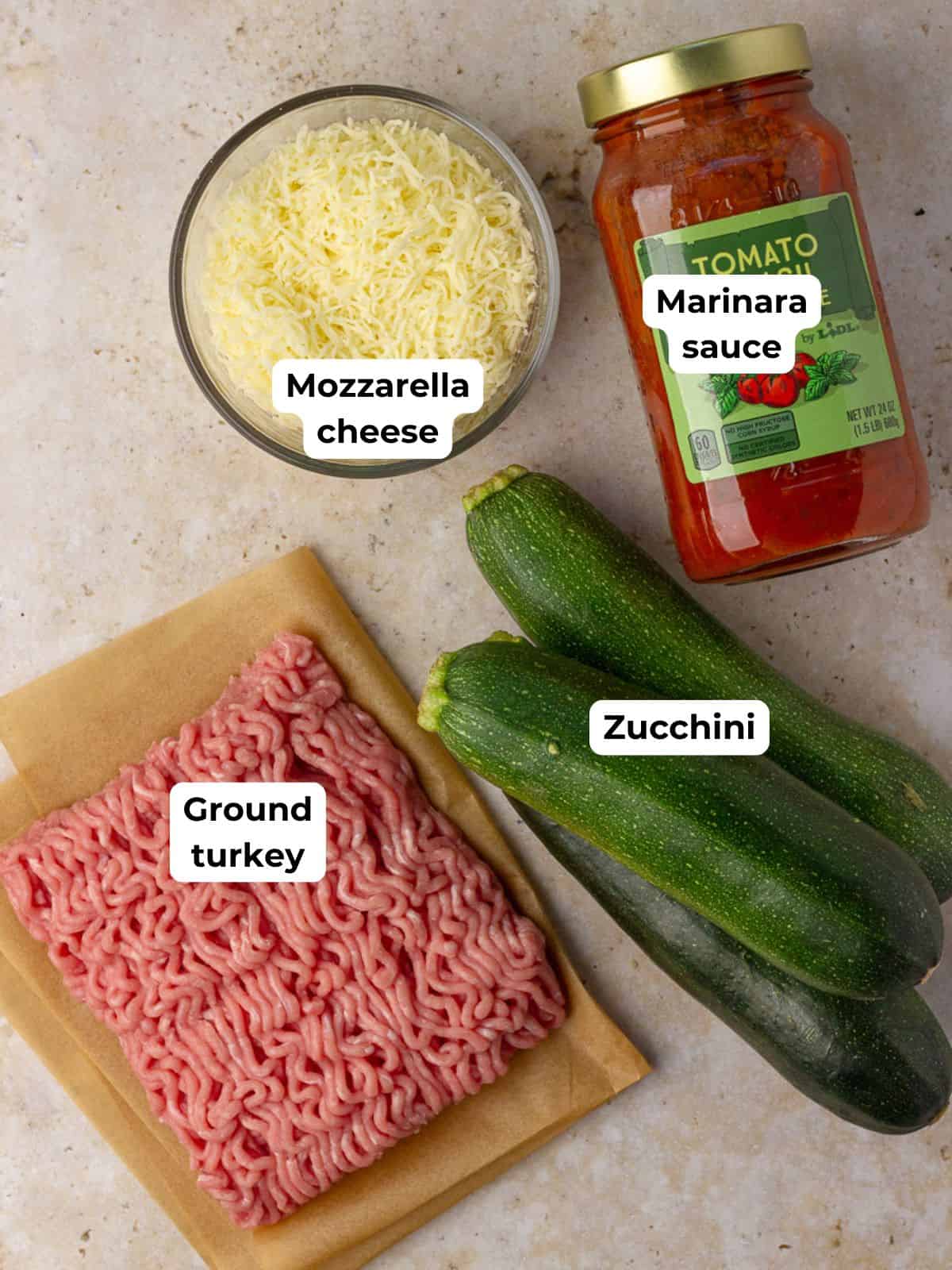 Labeled ingredients for zucchini boats: mozzarella cheese, marinara sauce, zucchini, and ground turkey arranged on a countertop.