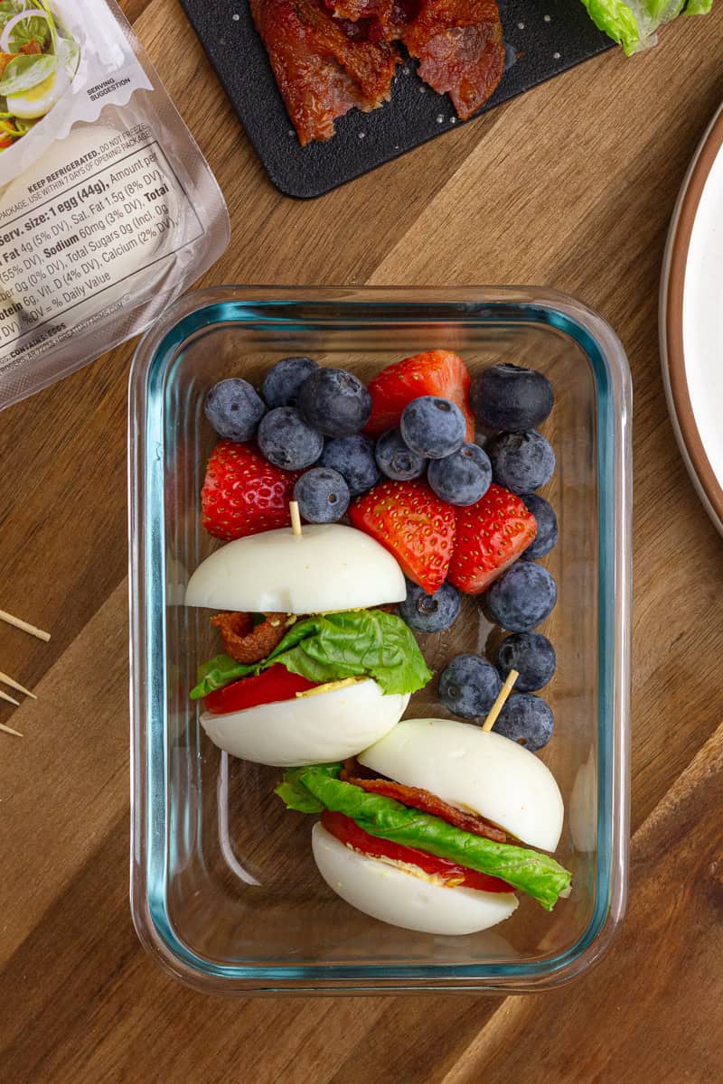 Two BLT egg sliders packed in a glass container with fresh blueberries and strawberries for a balanced, grab-and-go lunch.