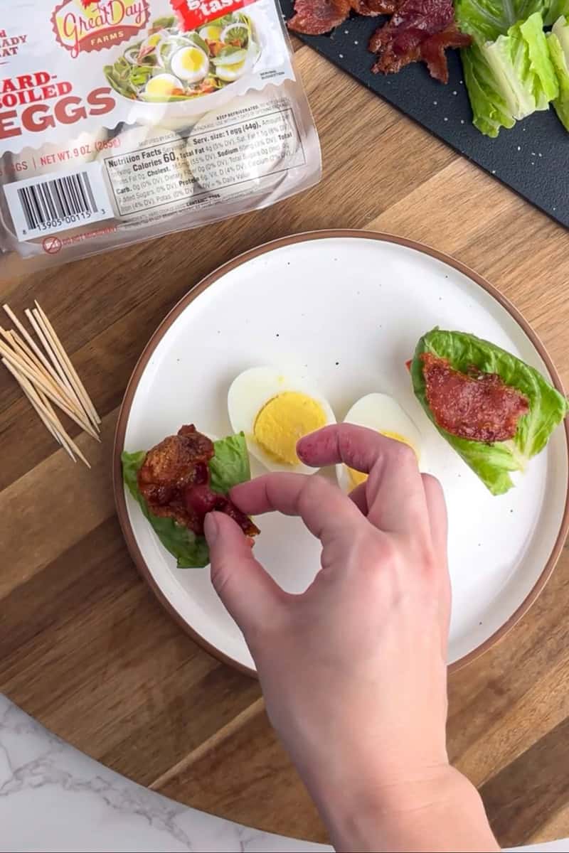 Layering crispy bacon on top of lettuce and tomato, ready to be sandwiched between egg halves.