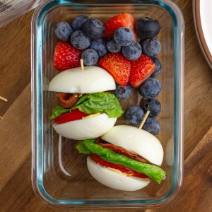 Glass meal prep container filled with two BLT egg sliders and a mix of blueberries and strawberries for a protein-packed snack box.