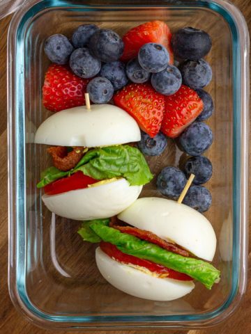 Glass meal prep container filled with two BLT egg sliders and a mix of blueberries and strawberries for a protein-packed snack box.