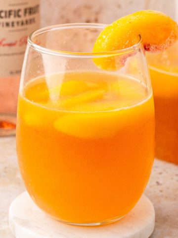 A stemless wine glass filled with bright orange peach sangria, garnished with a frozen peach slice.