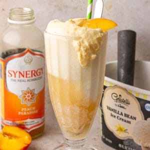 A tall glass filled with a peach kombucha float topped with a peach slice, with ice cream and kombucha in the background.
