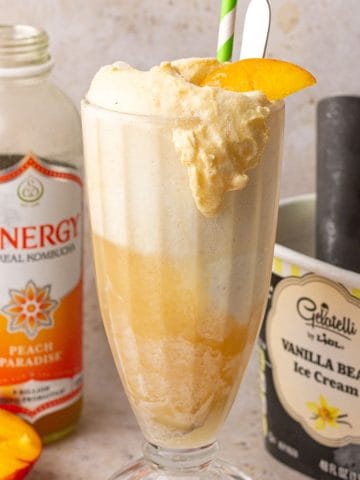 A tall glass filled with a peach kombucha float topped with a peach slice, with ice cream and kombucha in the background.