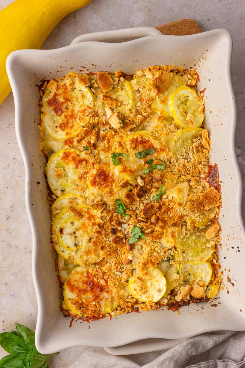 Freshly baked squash casserole in a white baking dish with a crisp topping and a yellow squash beside it.