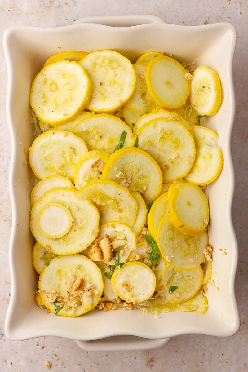 Unbaked yellow squash slices layered with basil and crumbs in a white baking dish, ready to go in the oven.