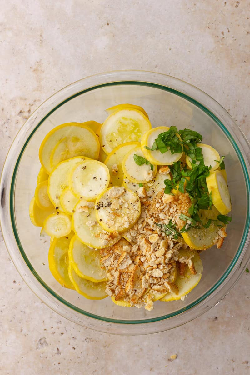A mixing bowl filled with sliced yellow squash, crushed crackers, basil, salt, and pepper.
