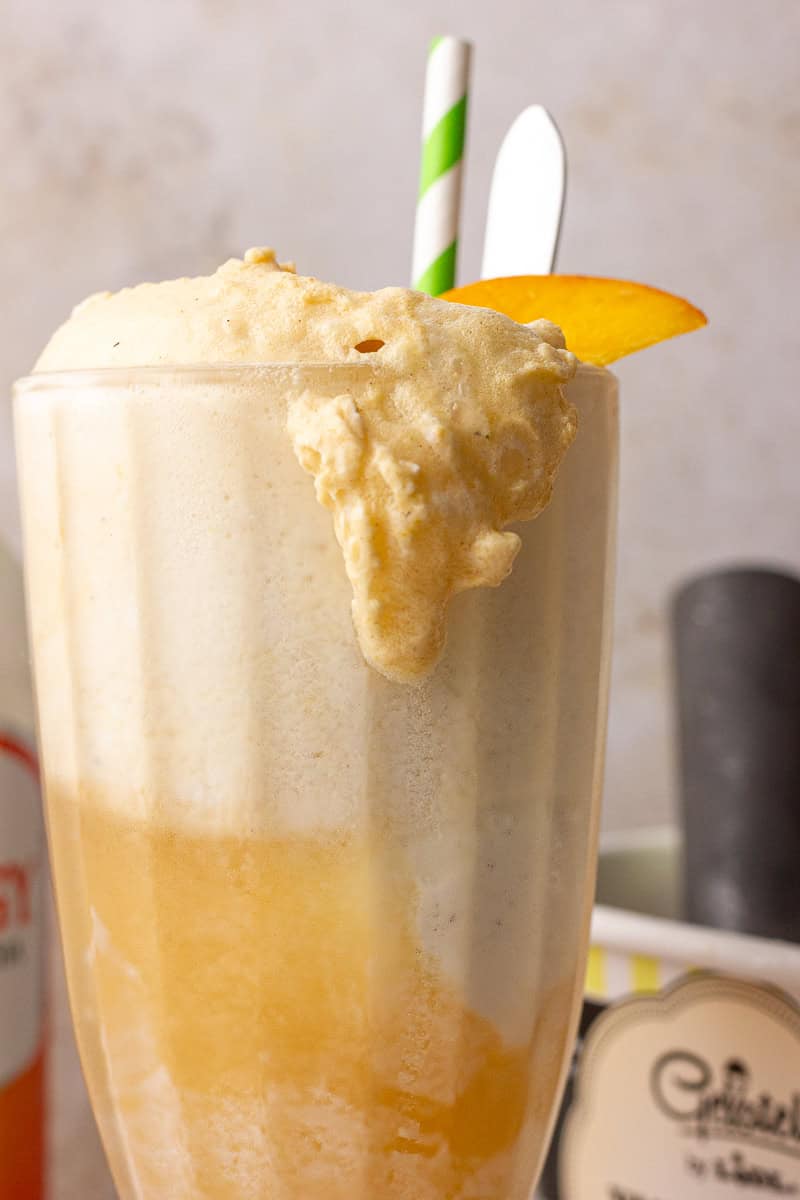 A close-up of the finished float with a peach wedge garnish and creamy foam spilling over the rim.