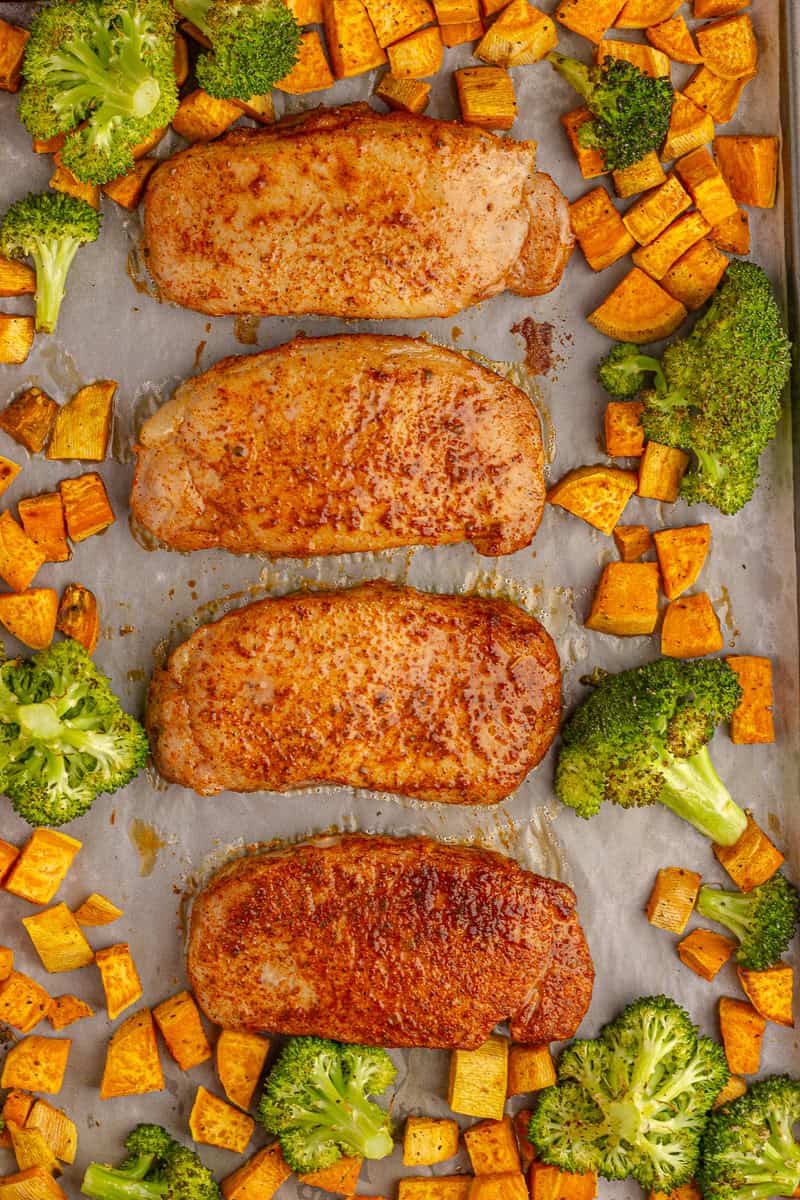 Cooked pork chops on a sheet pan surrounded by roasted sweet potatoes and broccoli.