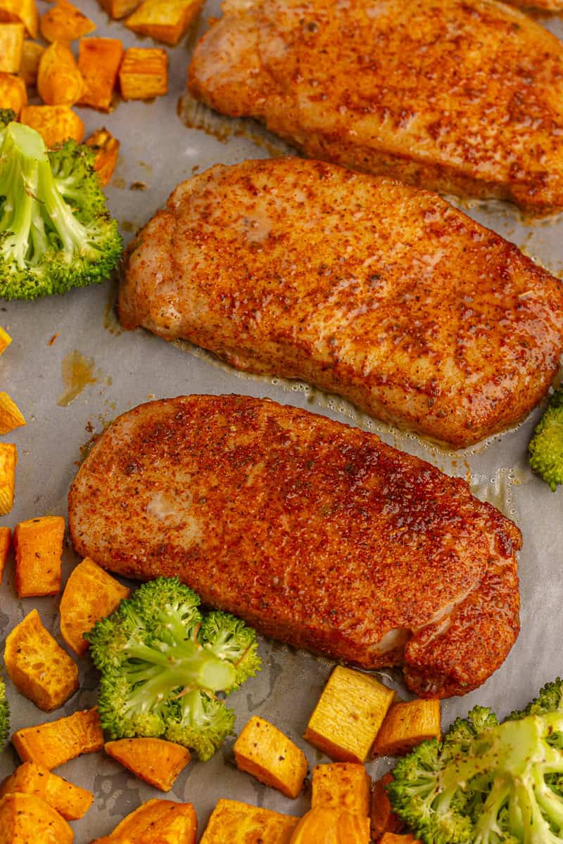 Close-up of roasted pork chops with golden seasoning, surrounded by broccoli and sweet potato cubes on a sheet pan.