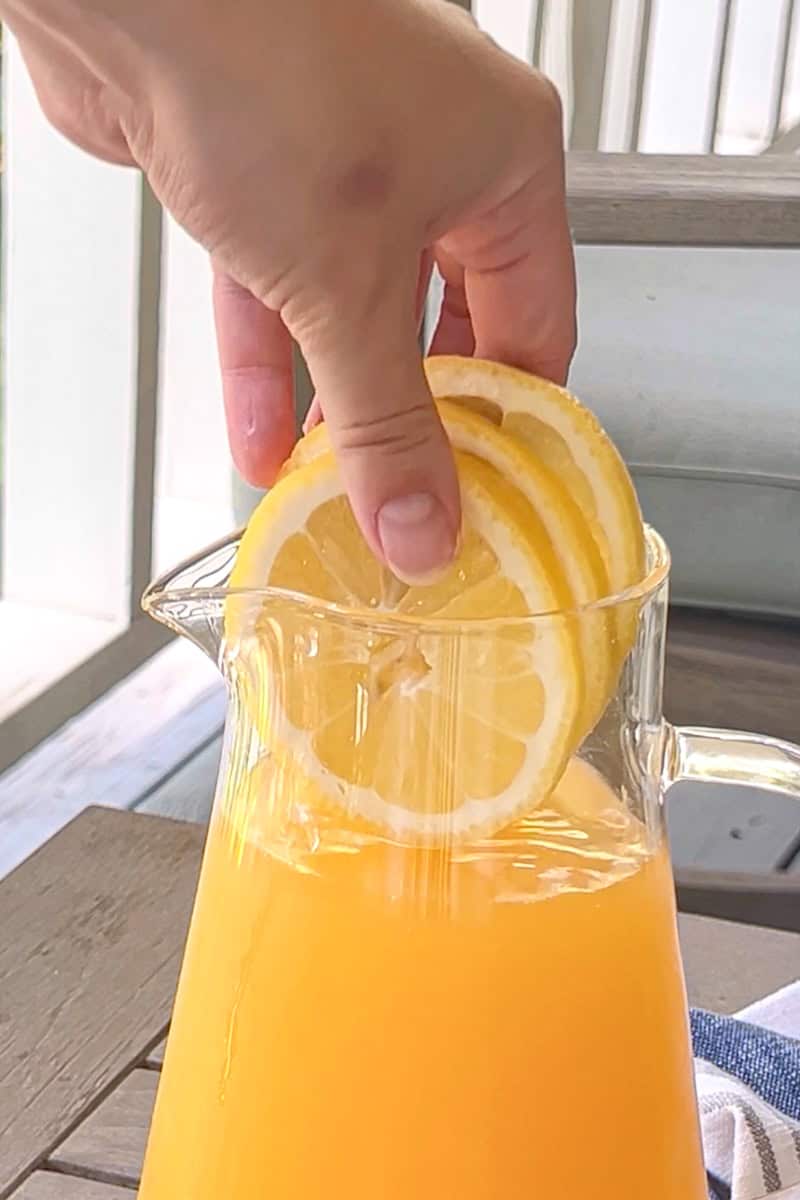 A hand placing fresh lemon slices into a clear pitcher of peach lemonade.