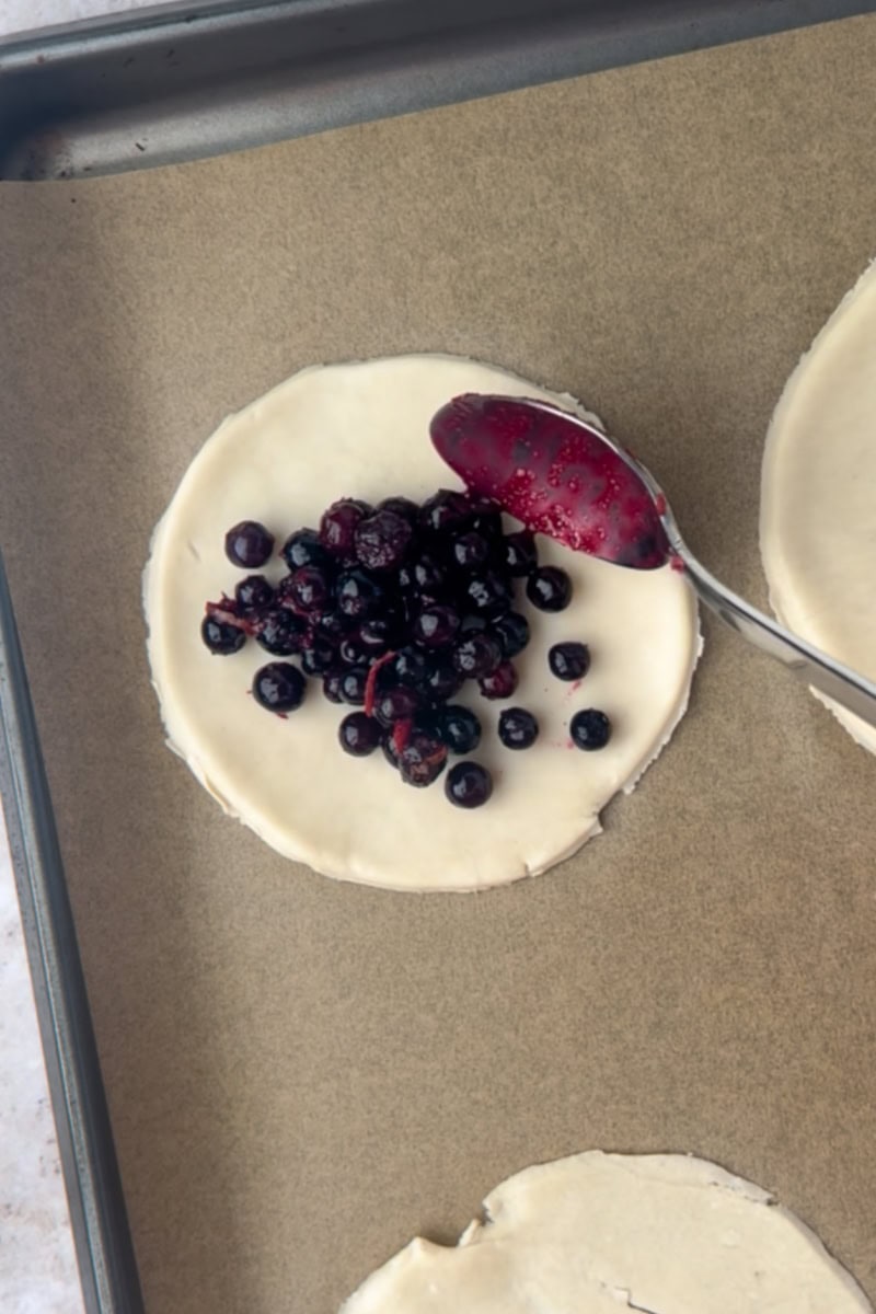 A spoon piles blueberry filling onto the center of a round pie-dough circle on a parchment-lined sheet.