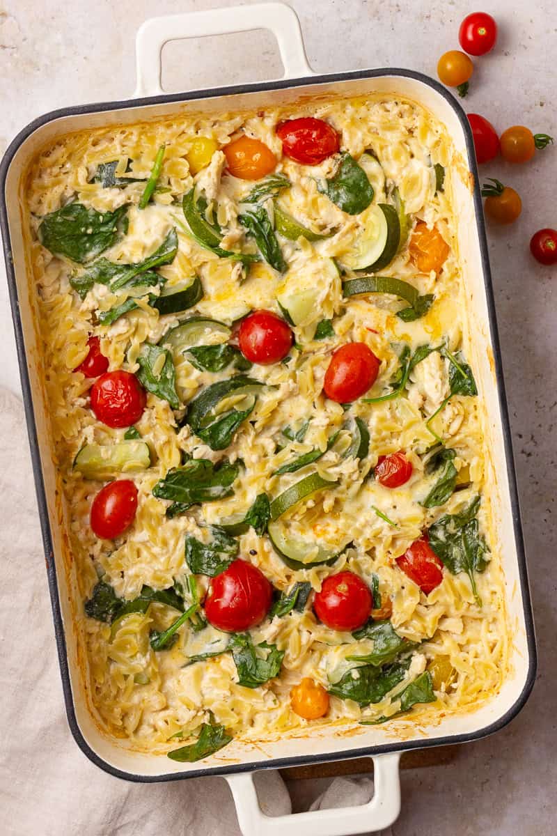 Golden baked Boursin chicken orzo in a white dish topped with spinach, zucchini, and cherry tomatoes.