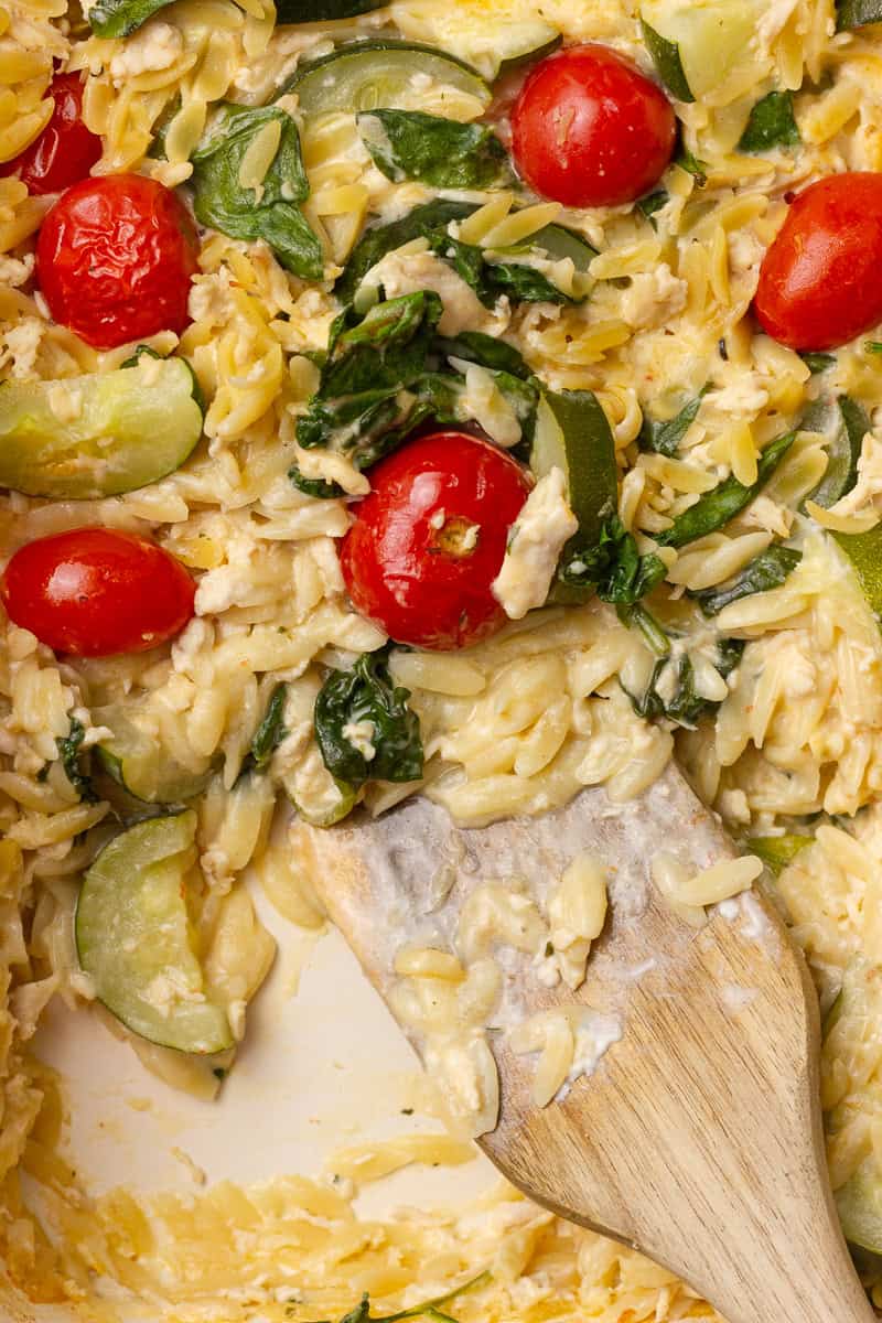Close-up of creamy Boursin chicken orzo with wilted spinach and juicy tomatoes, stirred with a wooden spoon.