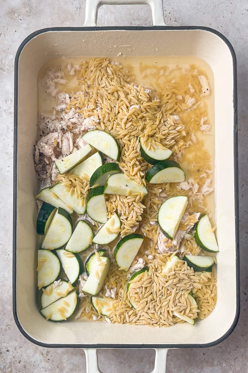 Unbaked chicken orzo casserole with zucchini, orzo, and broth layered in a cream-colored baking dish.