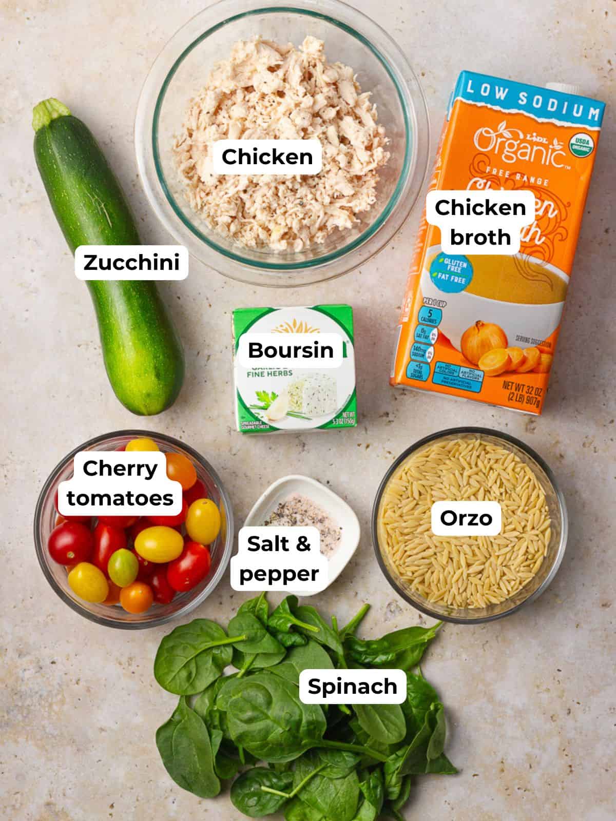 Flat lay of labeled ingredients for Boursin chicken orzo including orzo, spinach, zucchini, cherry tomatoes, broth, and cheese.