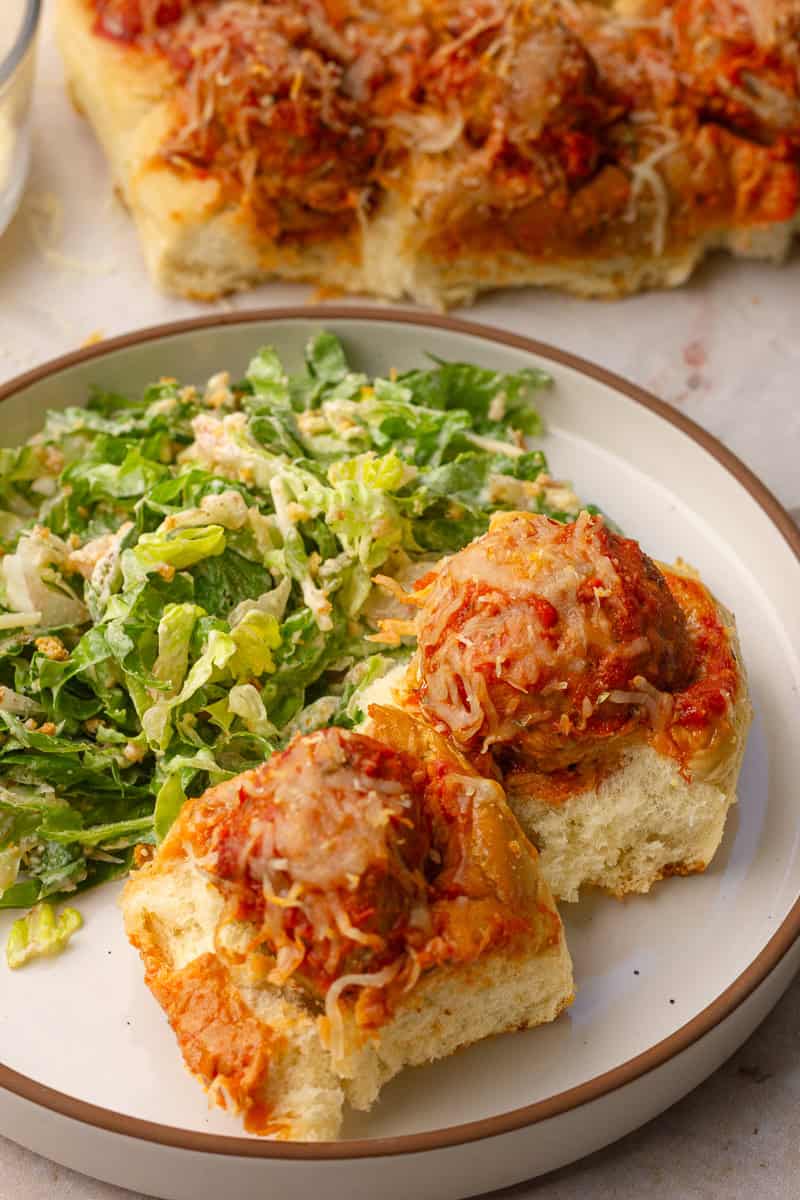 Two meatball sliders on a plate with chopped Caesar salad, served alongside the rest of the pull-apart sliders in the background.