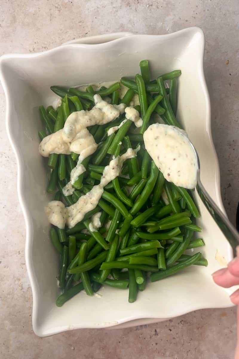 Green beans in a white casserole dish being topped with spoonfuls of creamy sauce.