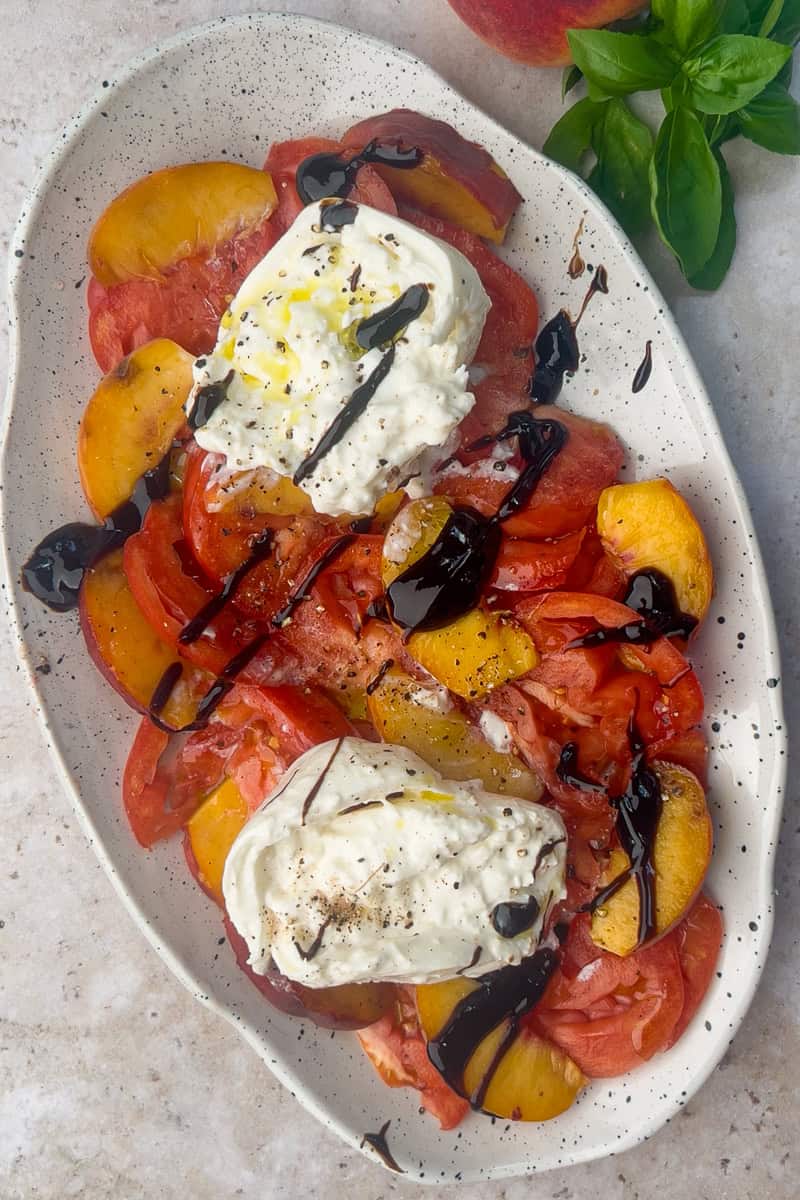 Finished salad with balsamic glaze drizzled over burrata, tomatoes, and peaches on a white platter.