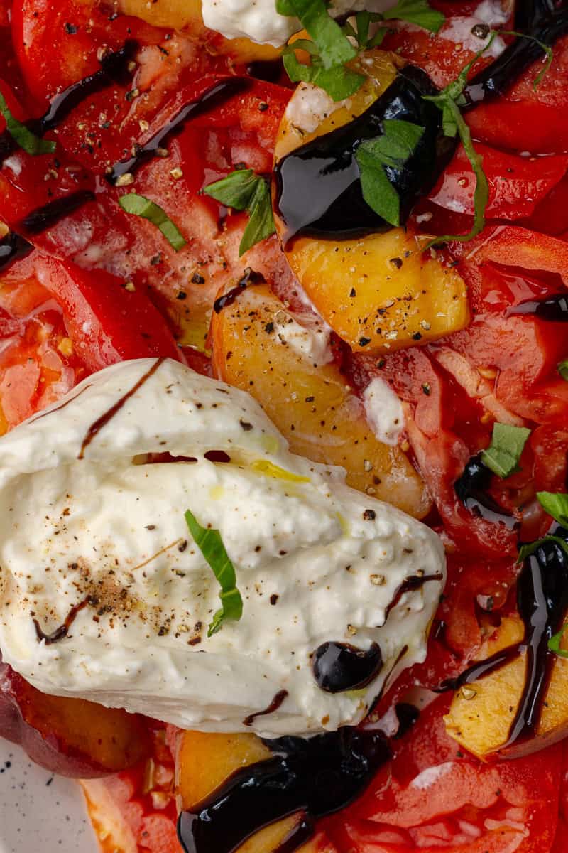 Close-up of creamy burrata, juicy peaches, and tomatoes drizzled with balsamic glaze and topped with fresh basil.