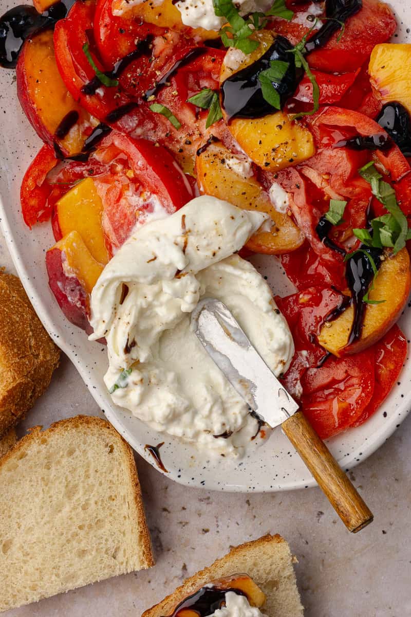 Partially eaten peach tomato burrata salad with a knife spread into the creamy burrata and slices of bread nearby.