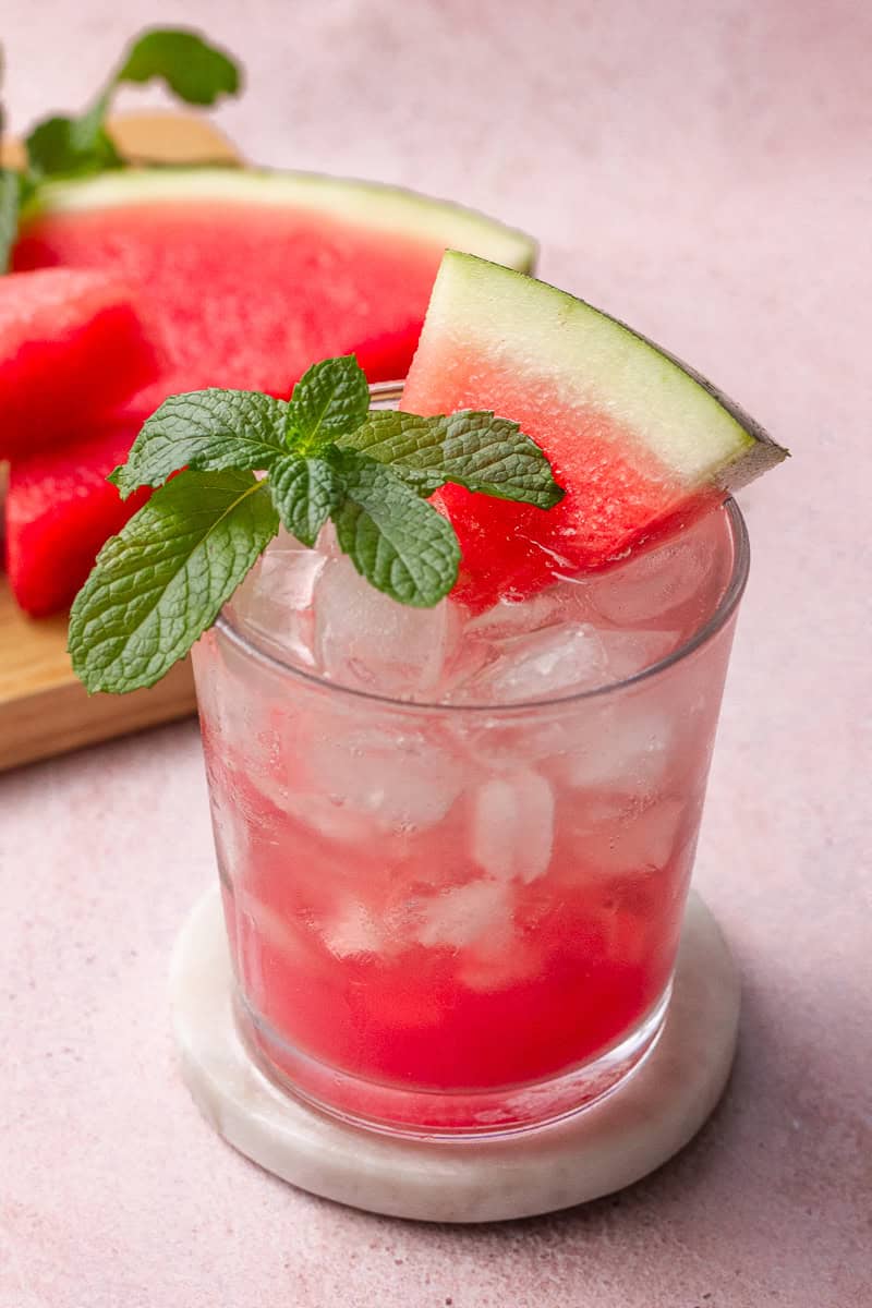 A glass of virgin watermelon mojito with mint and a watermelon slice, styled on a pink surface with fresh watermelon in the background.