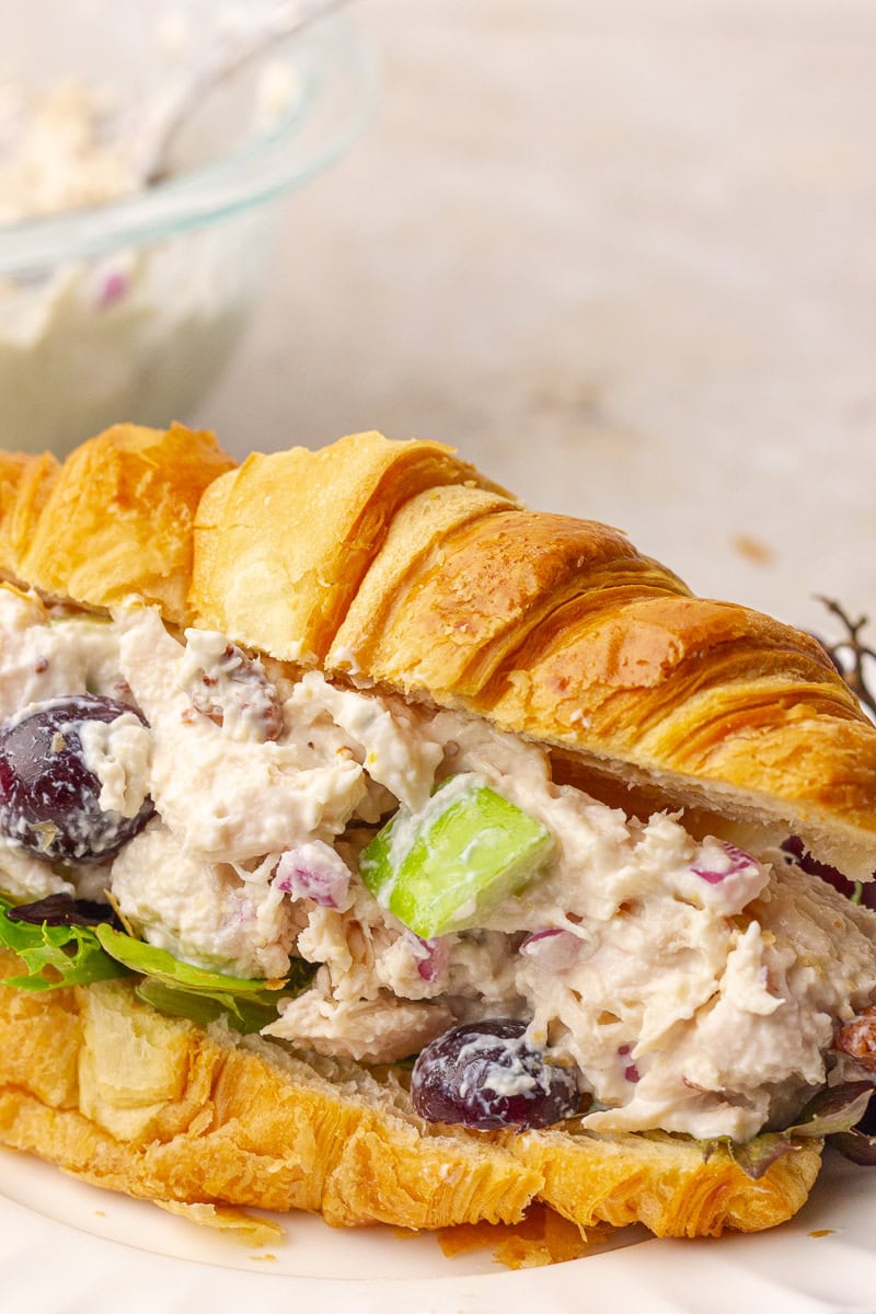 Front view of a flaky croissant stuffed with creamy chicken salad, showing grapes, apples, and greens.
