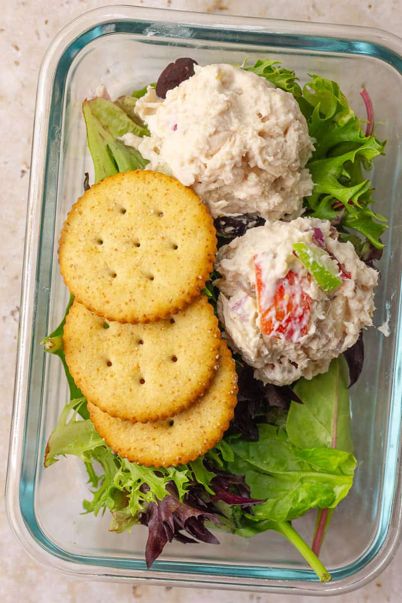 Meal prep container with scoops of chicken salad over greens and a side of round crackers.