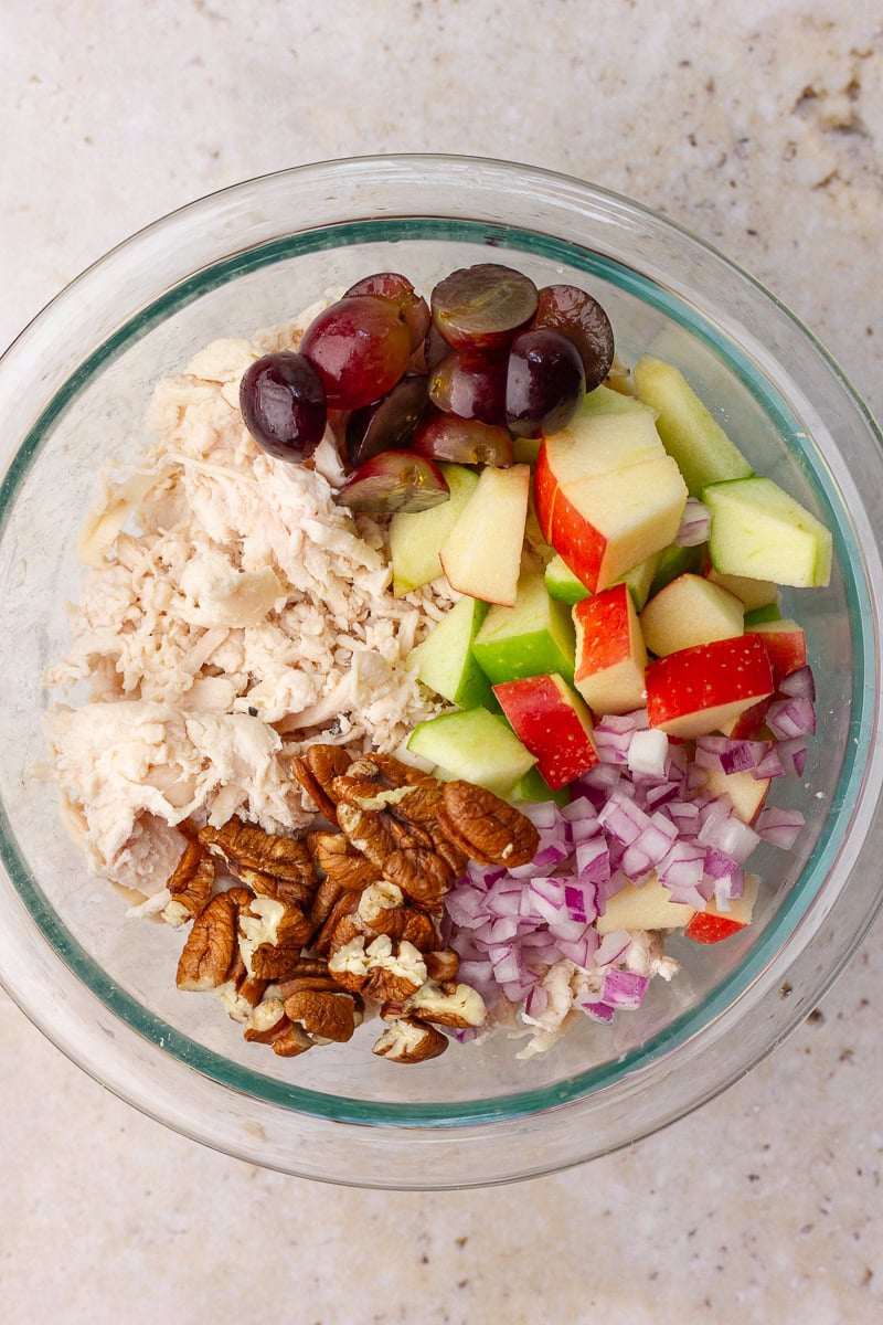 Bowl of salad ingredients before mixing, including apples, grapes, pecans, red onion, and shredded chicken.