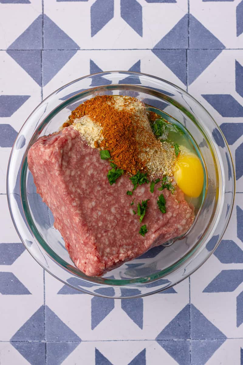 Ground lamb, egg, parsley, breadcrumbs, and gyro seasoning are shown in a glass bowl before mixing.