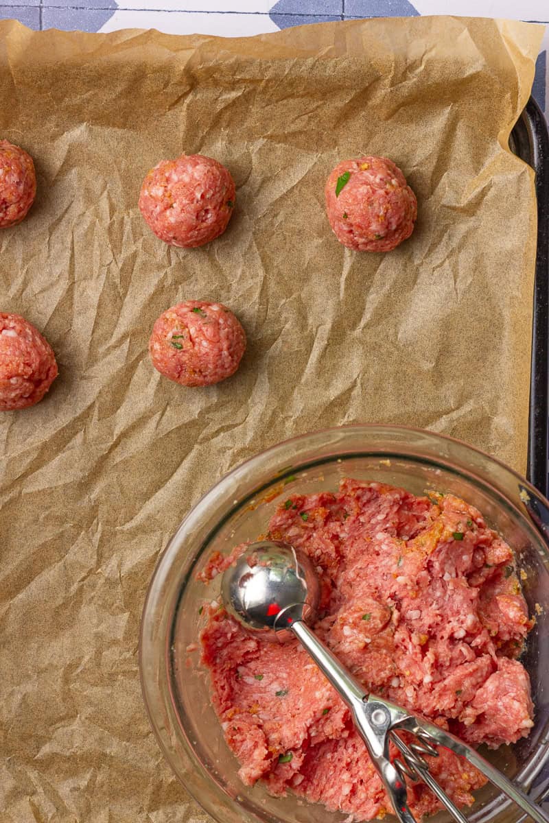 Scooped lamb meatball mixture is shown in a bowl with a cookie scoop, and shaped meatballs are arranged on a parchment-lined baking sheet.