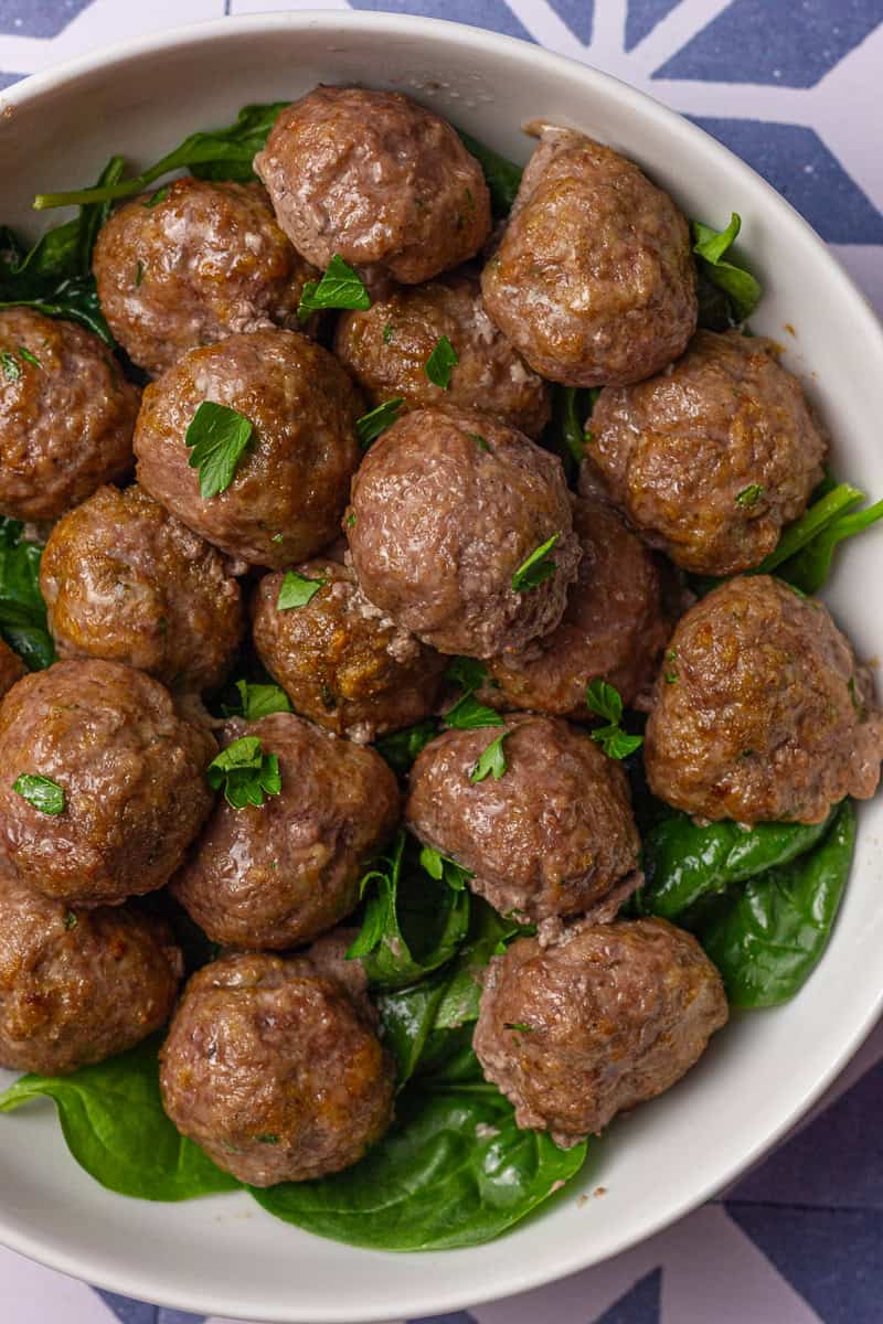 Close-up of juicy cooked lamb meatballs on a bed of spinach with fresh parsley on top, served in a white bowl.
