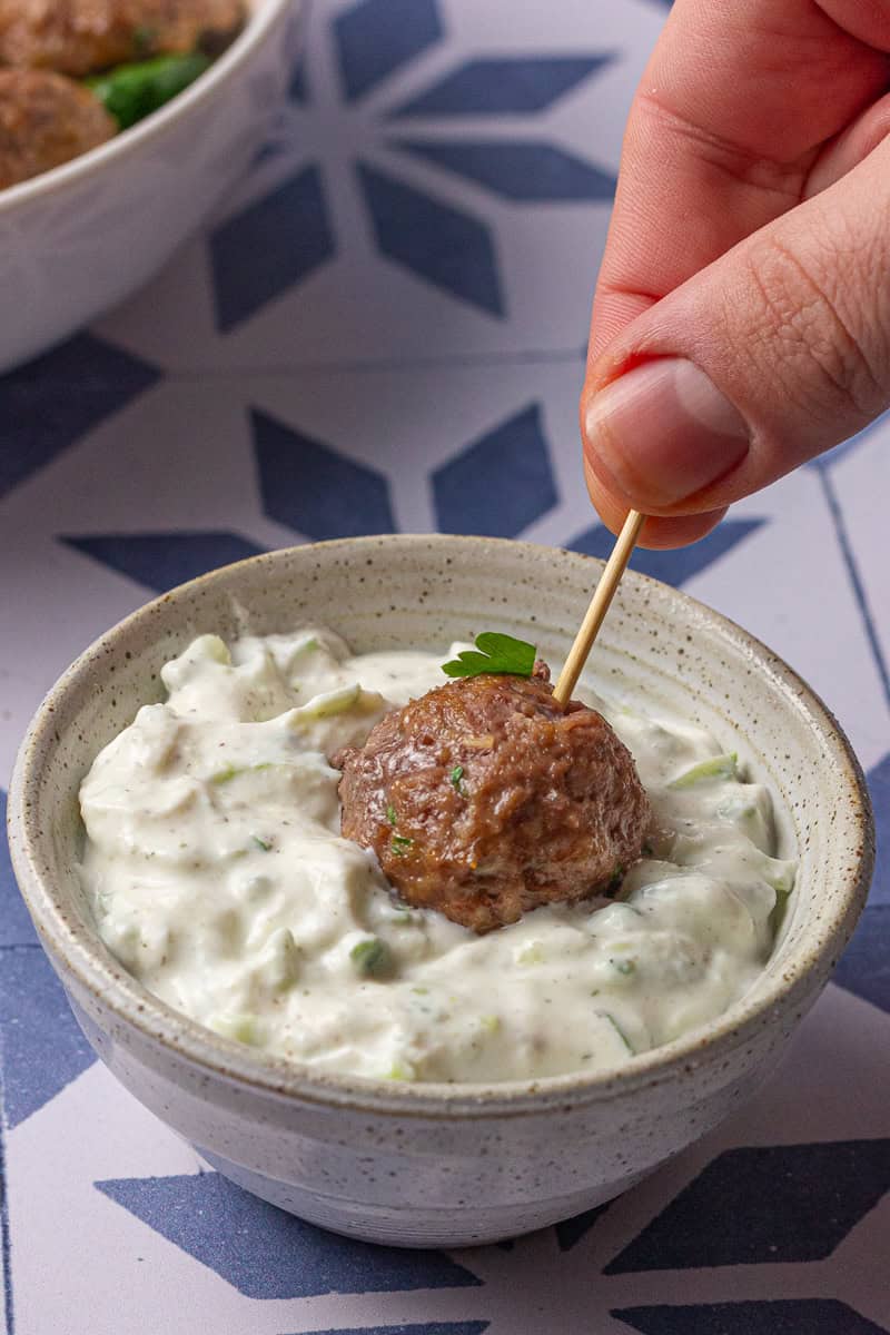 A toothpick holds a lamb meatball dipped in creamy tzatziki sauce in a small speckled bowl.