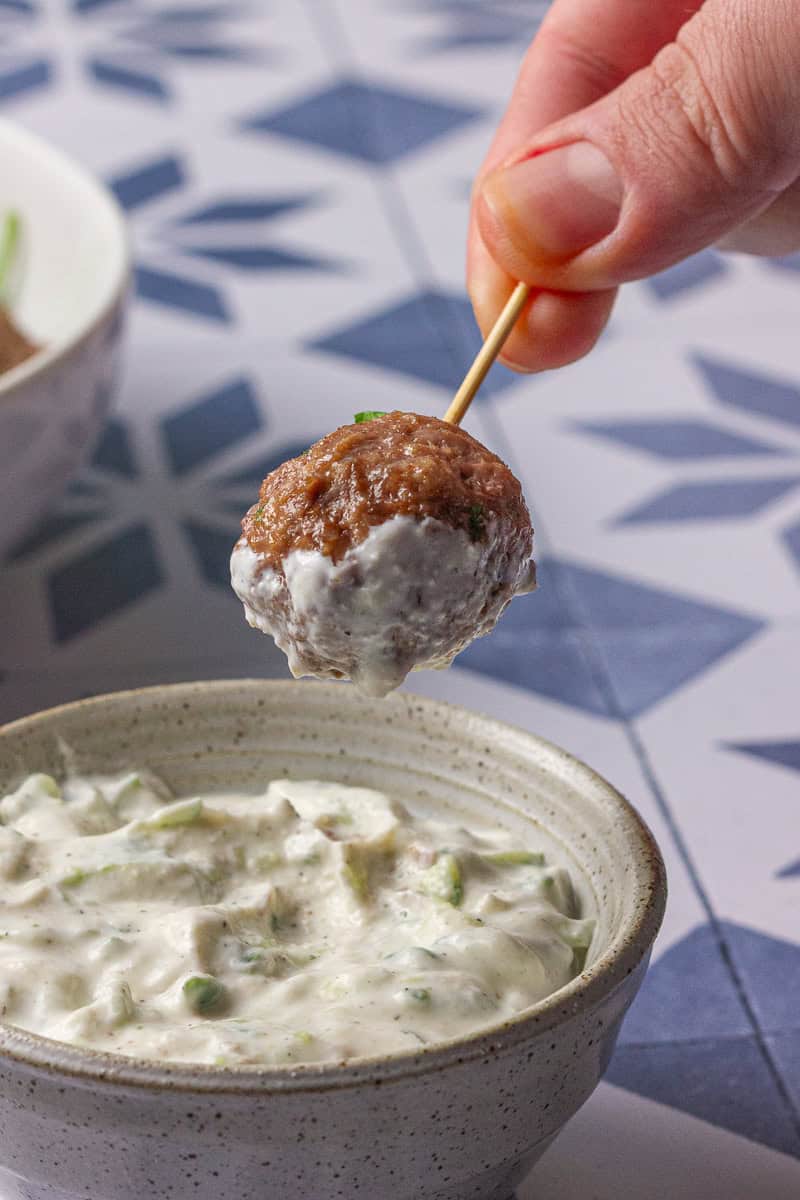 A single meatball is lifted from the sauce, showing a thick coating of tzatziki on top.